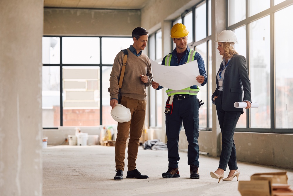 Three Construction Professionals Review Blueprints Inside a Partly Finished Building Inside a Bright Space