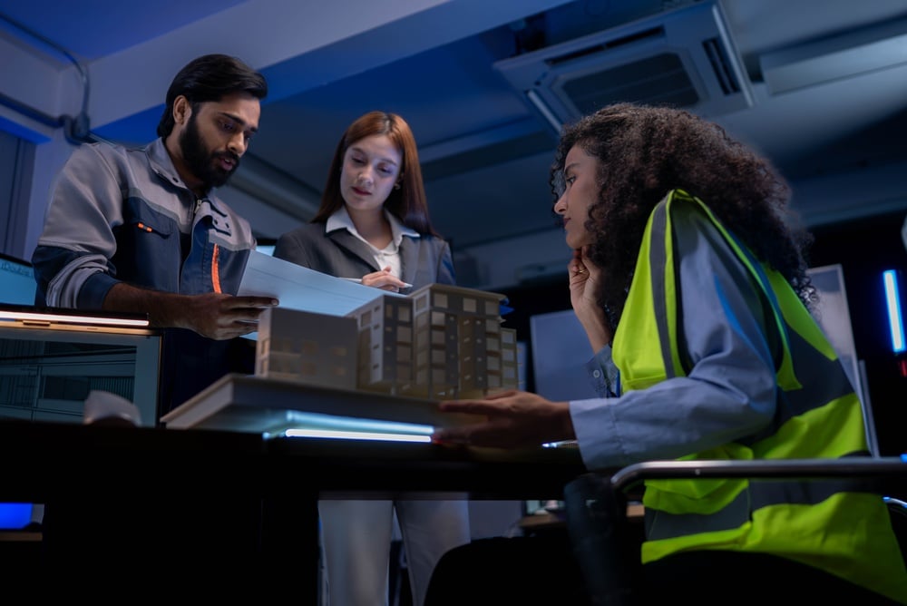 Three engineers review a building model and blueprints in a blue-lit lab setup.